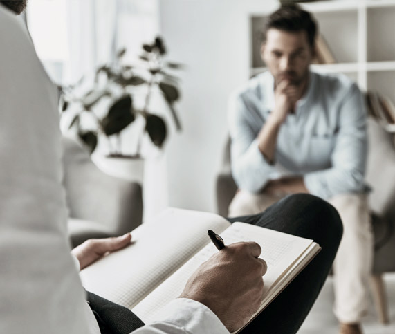Psychotherapy session in which two men are sat opposite each other in a therapy room with one taking notes on a clipboard